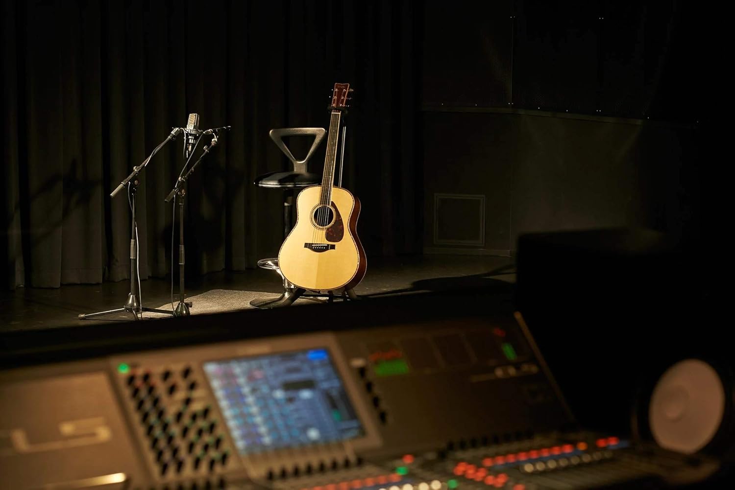 Cena de palco com violão Yamaha LS6 A.R.E. apoiado em cadeira preta com encosto triangular. Ao lado, dois microfones em pedestais. Fundo com cortinas pretas e ambiente de estúdio ou show