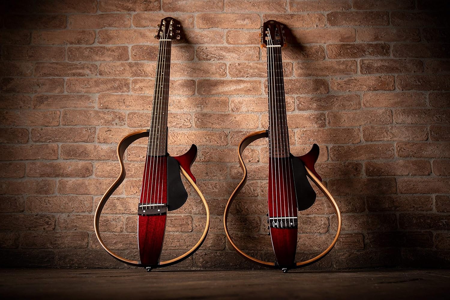 Dois violões Yamaha SLG-200N TBL com corpo esqueleto e acabamento translúcido vermelho em fundo de parede de tijolos.